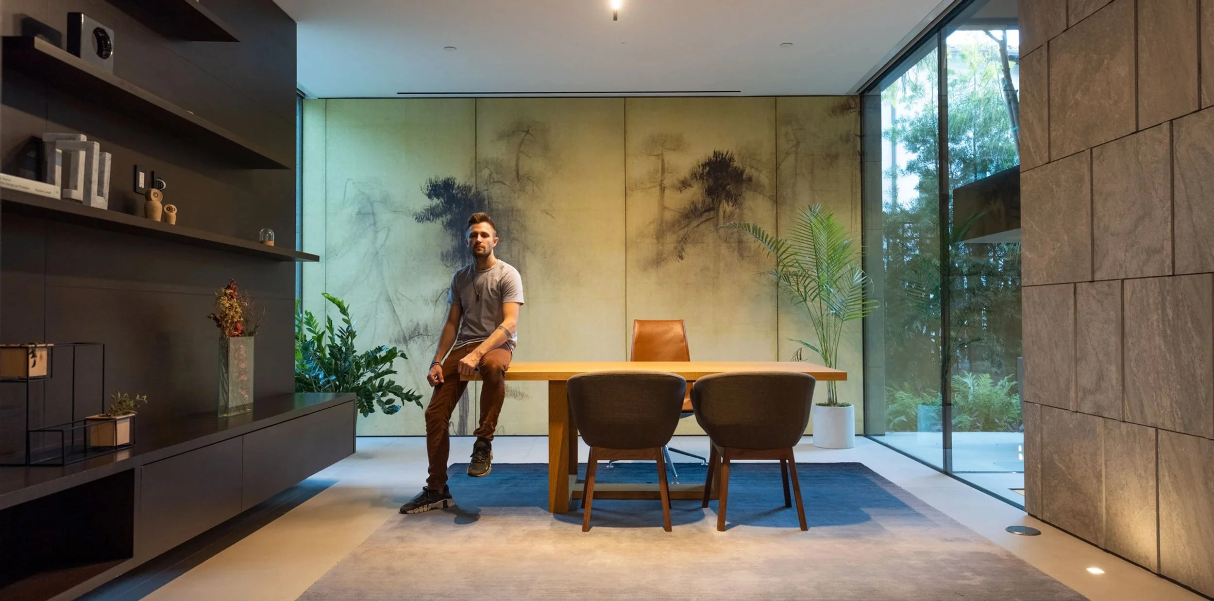 Joe Bryant seated on a large wood desk in a room with a landscape mural and garden window.