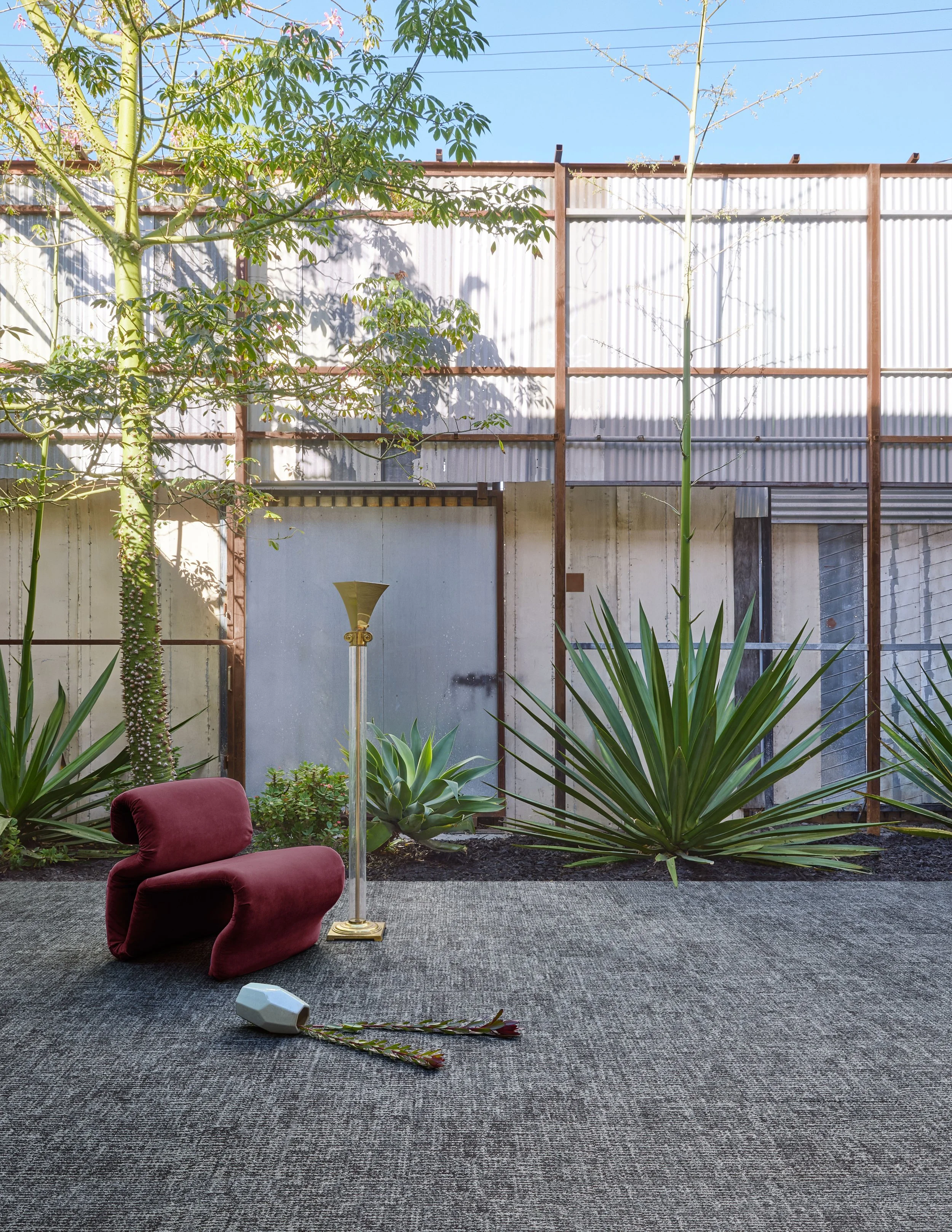 Red velvet lounge chair and gold lamp on gray carpet against corrugated metal in bright sunlight.