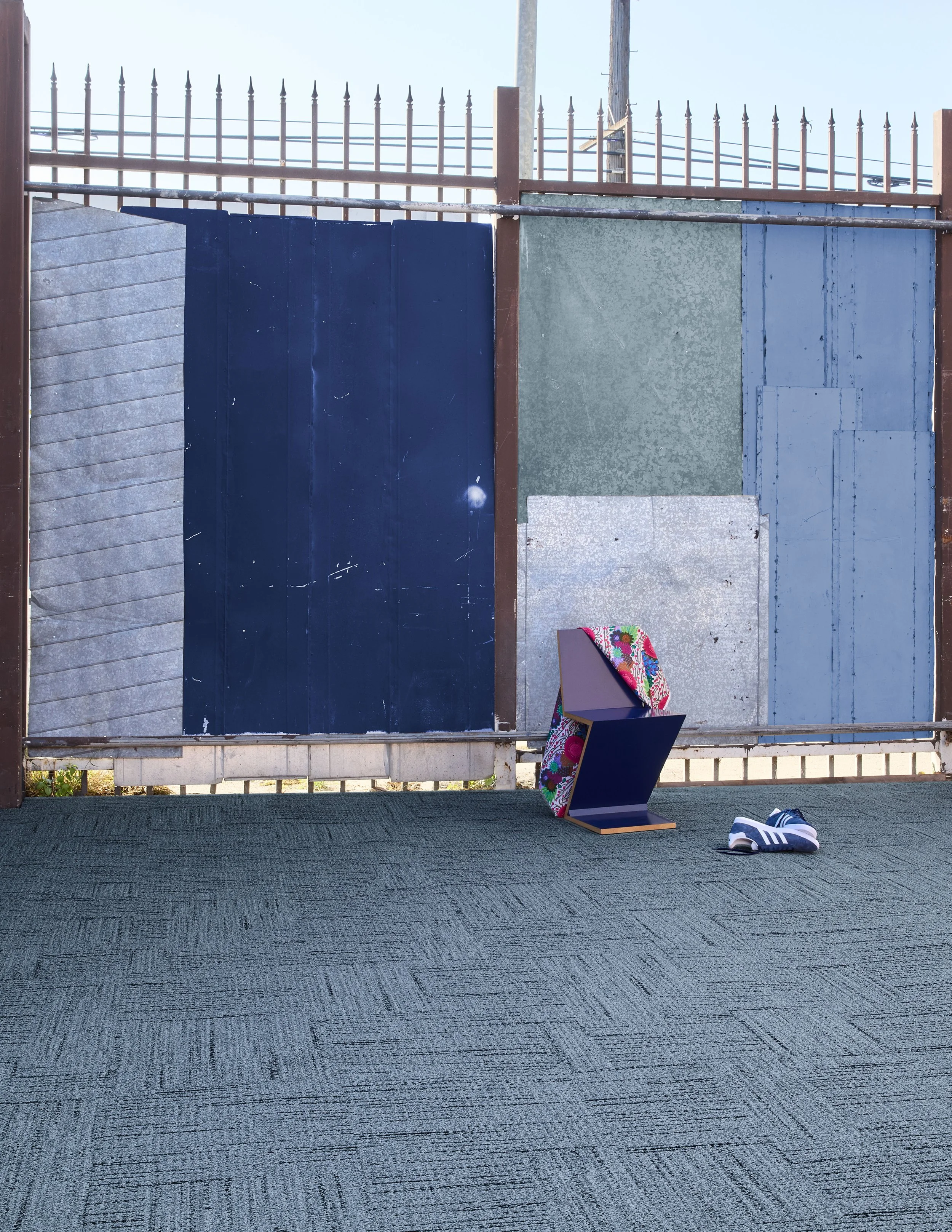 Dark blue geometric chair draped with floral fabric on gray carpet before an industrial metal fence.