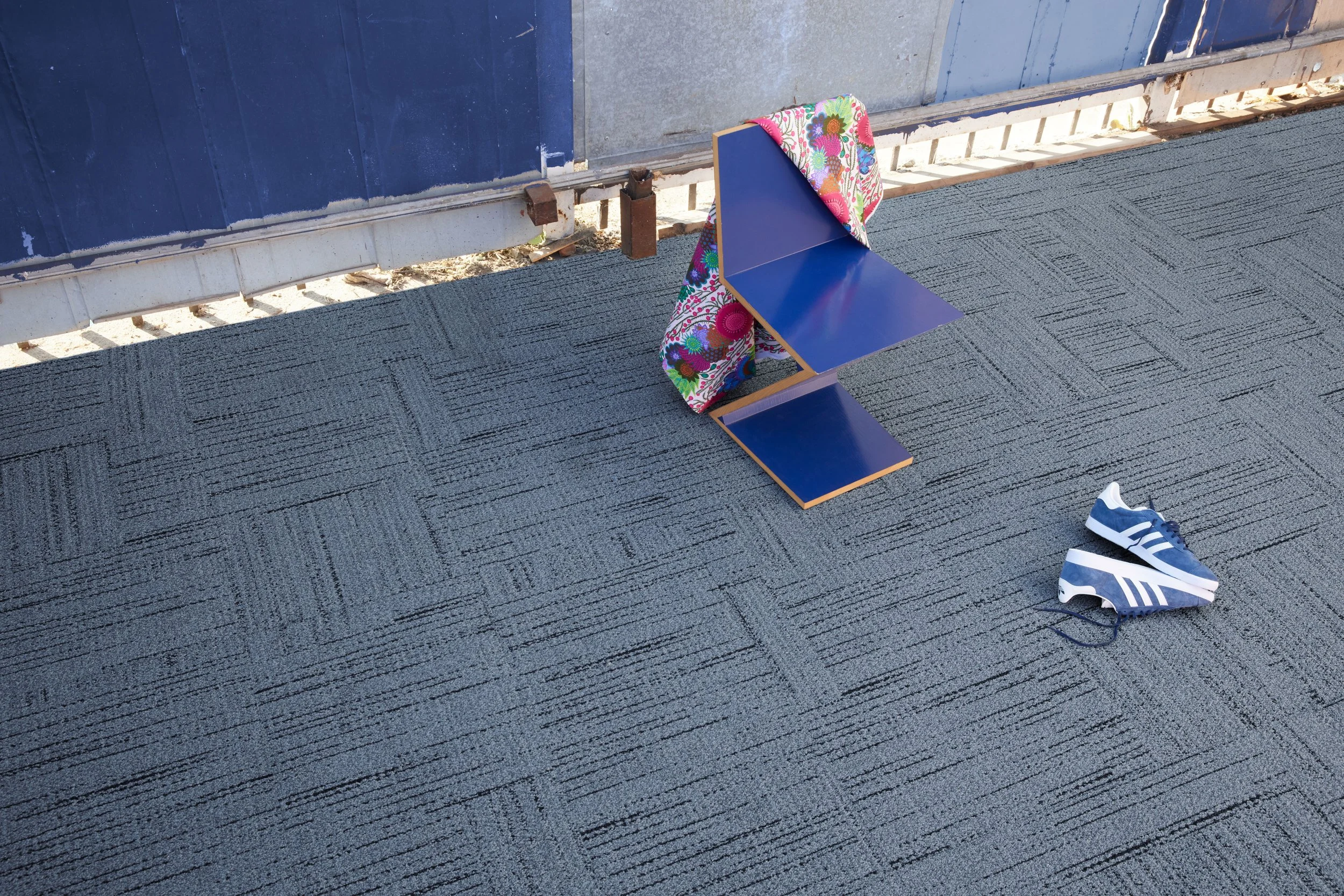 Blue zig-zag chair with a floral scarf and sneakers on gray carpet tiles in bright daylight.