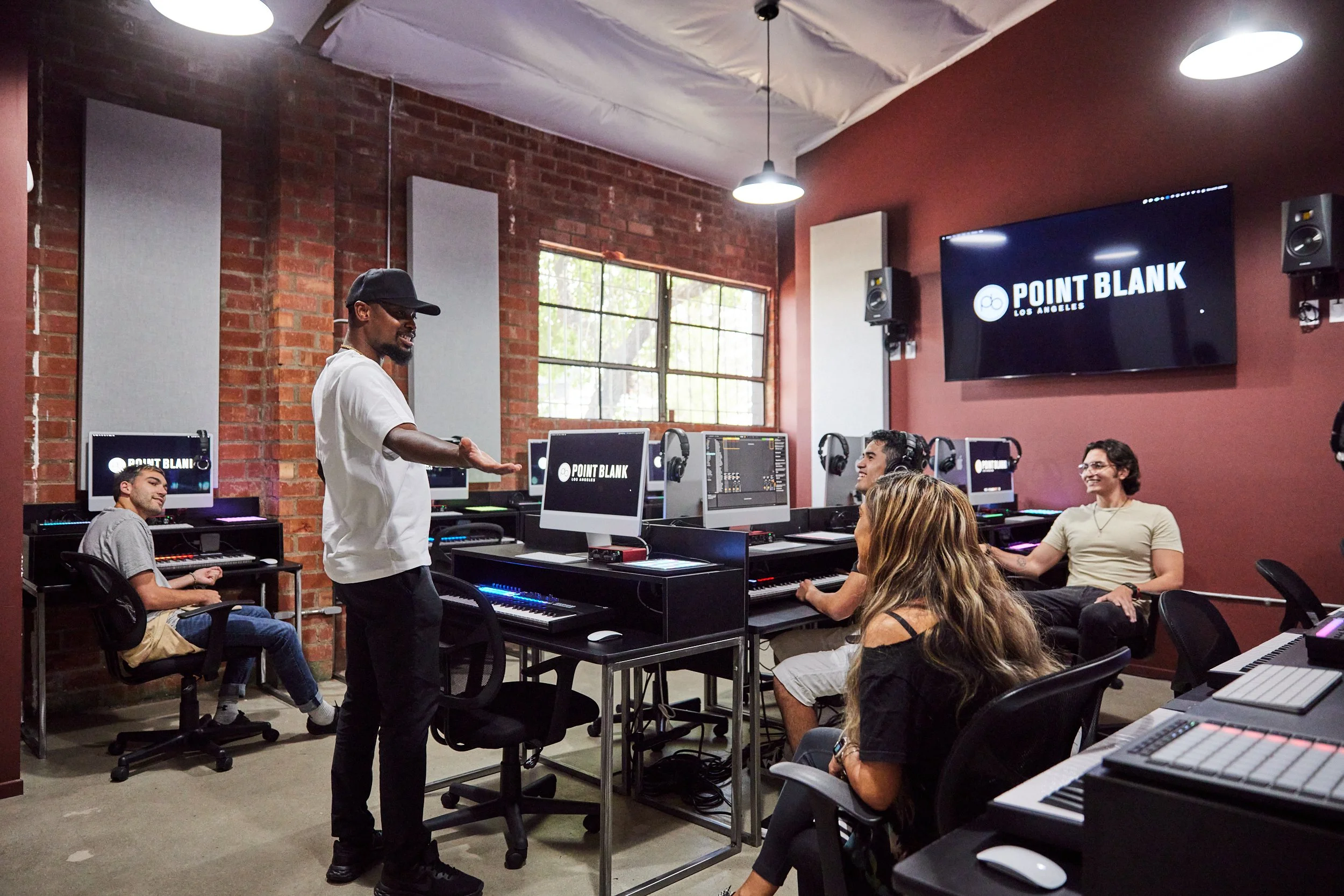 Instructor gesturing to students at workstations in a Point Blank studio with brick and acoustic panels.