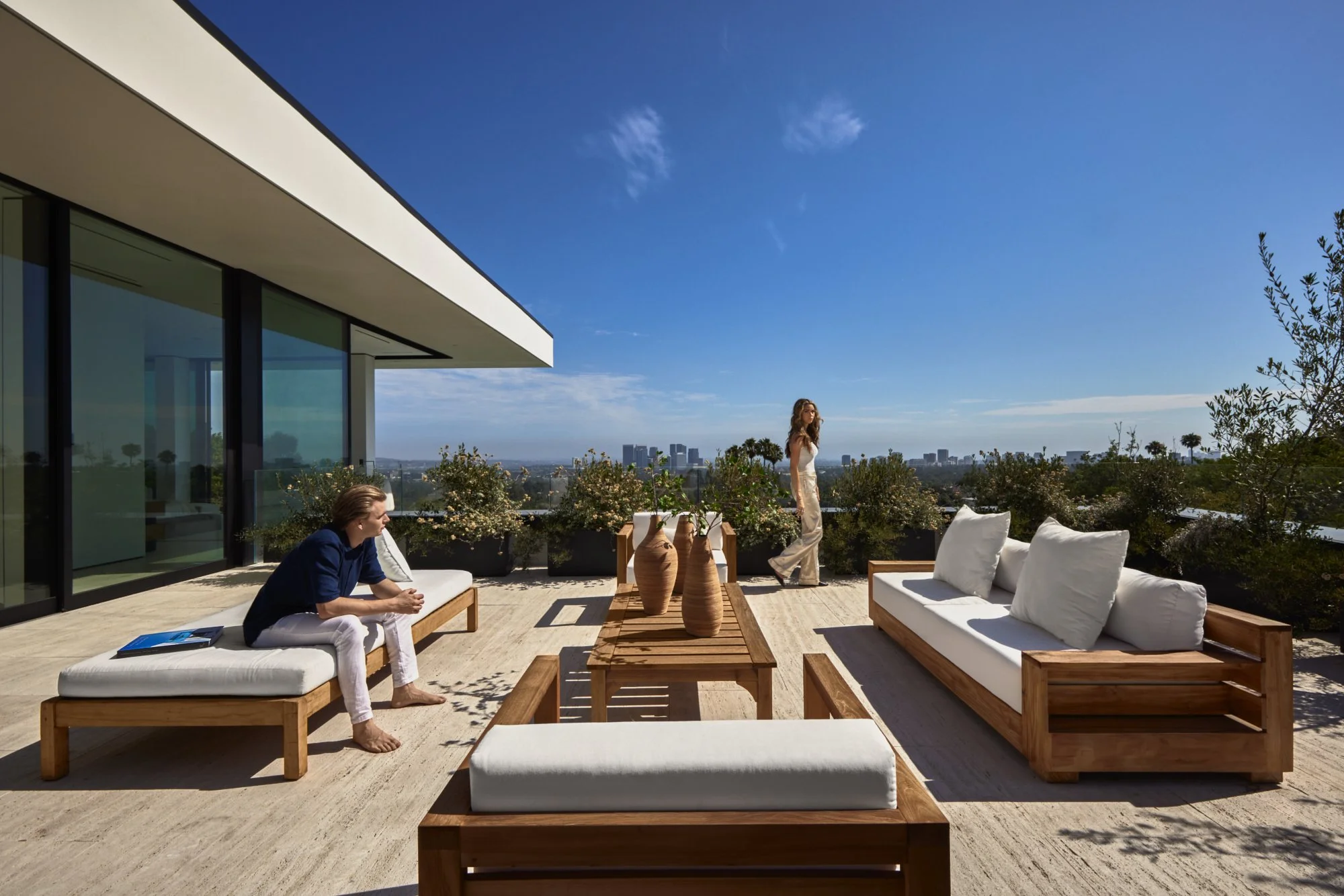 Sunlit terrace with wood lounge furniture and white cushions overlooking a distant city skyline.