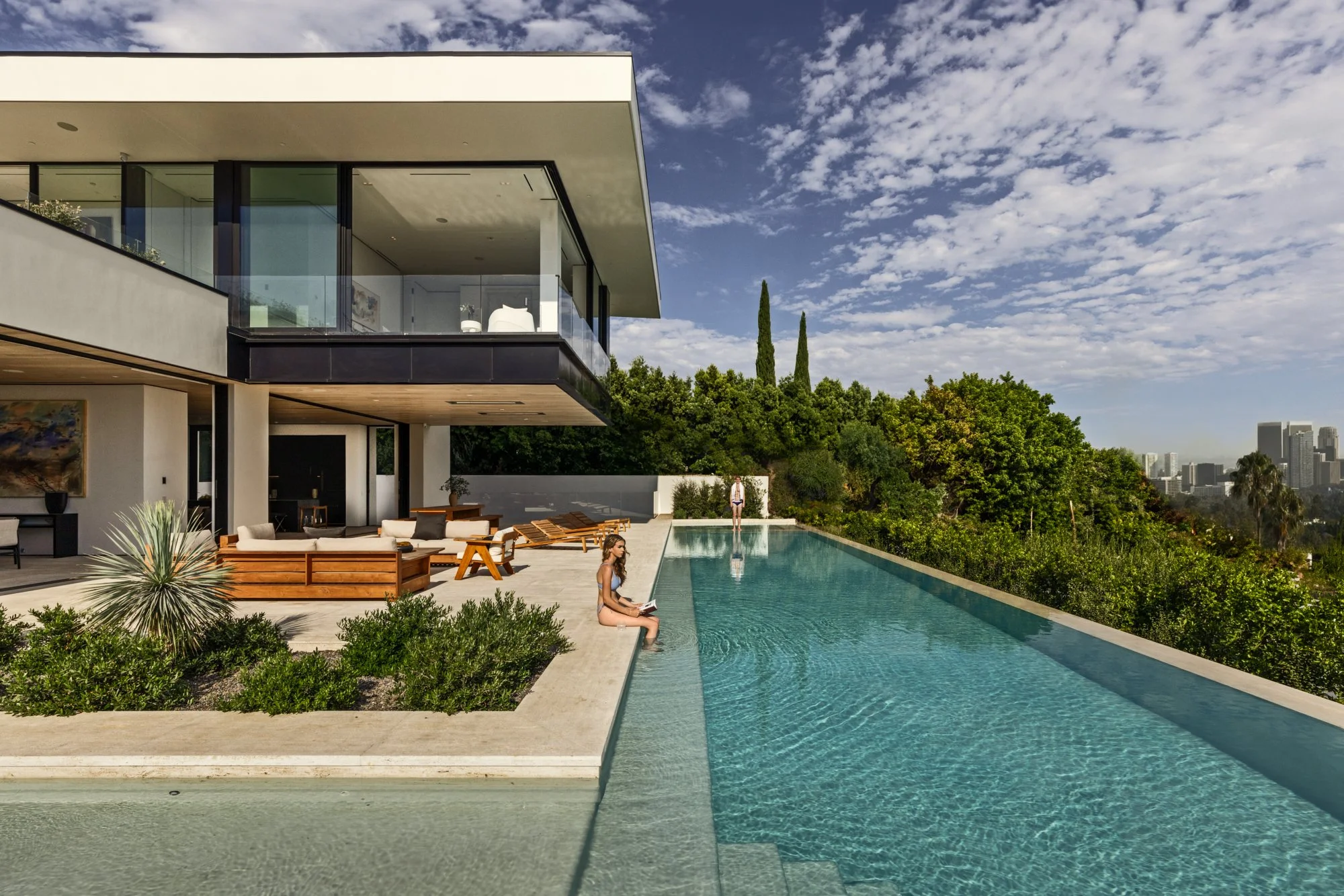Hillside white glass residence with a long lap pool and city skyline beyond under a bright sky.