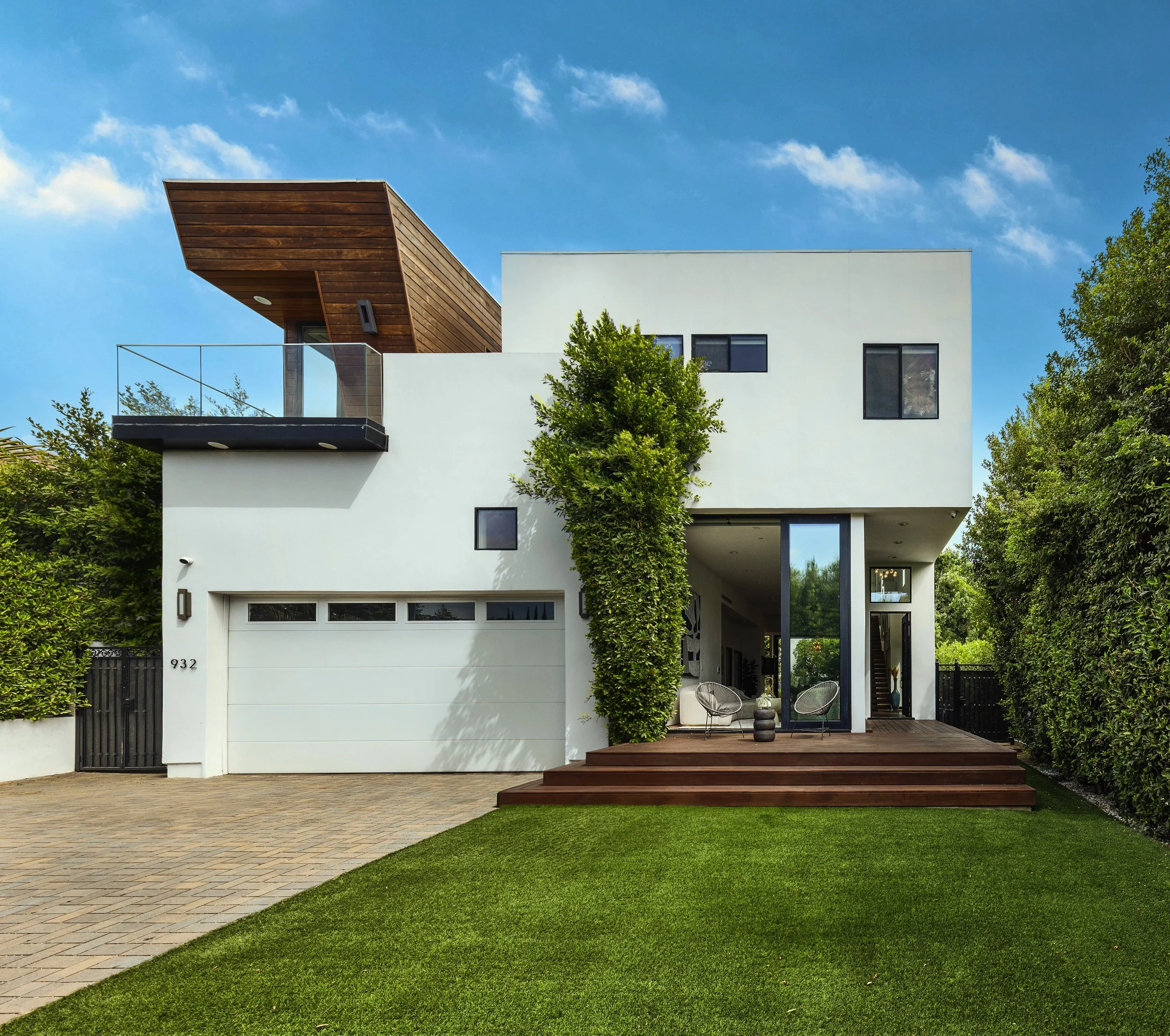White two-story home with a cantilevered wood balcony and manicured lawn in bright daylight.