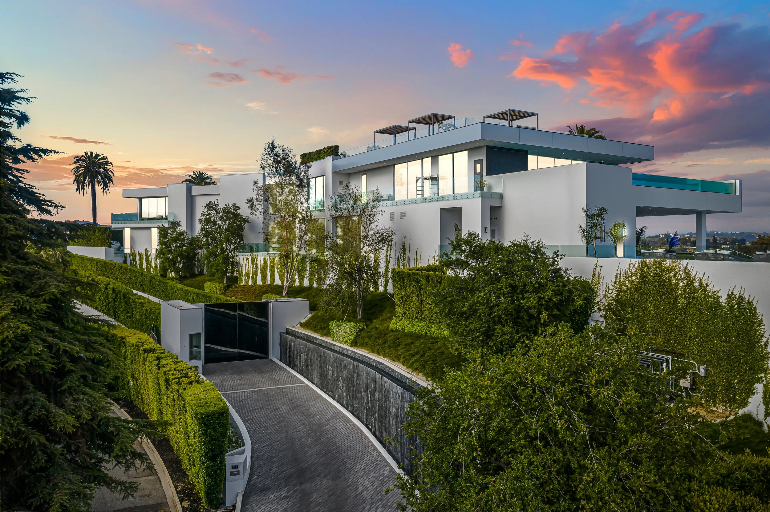 White multi-level hillside mansion at sunset with glass walls, terraces, and a curved driveway.