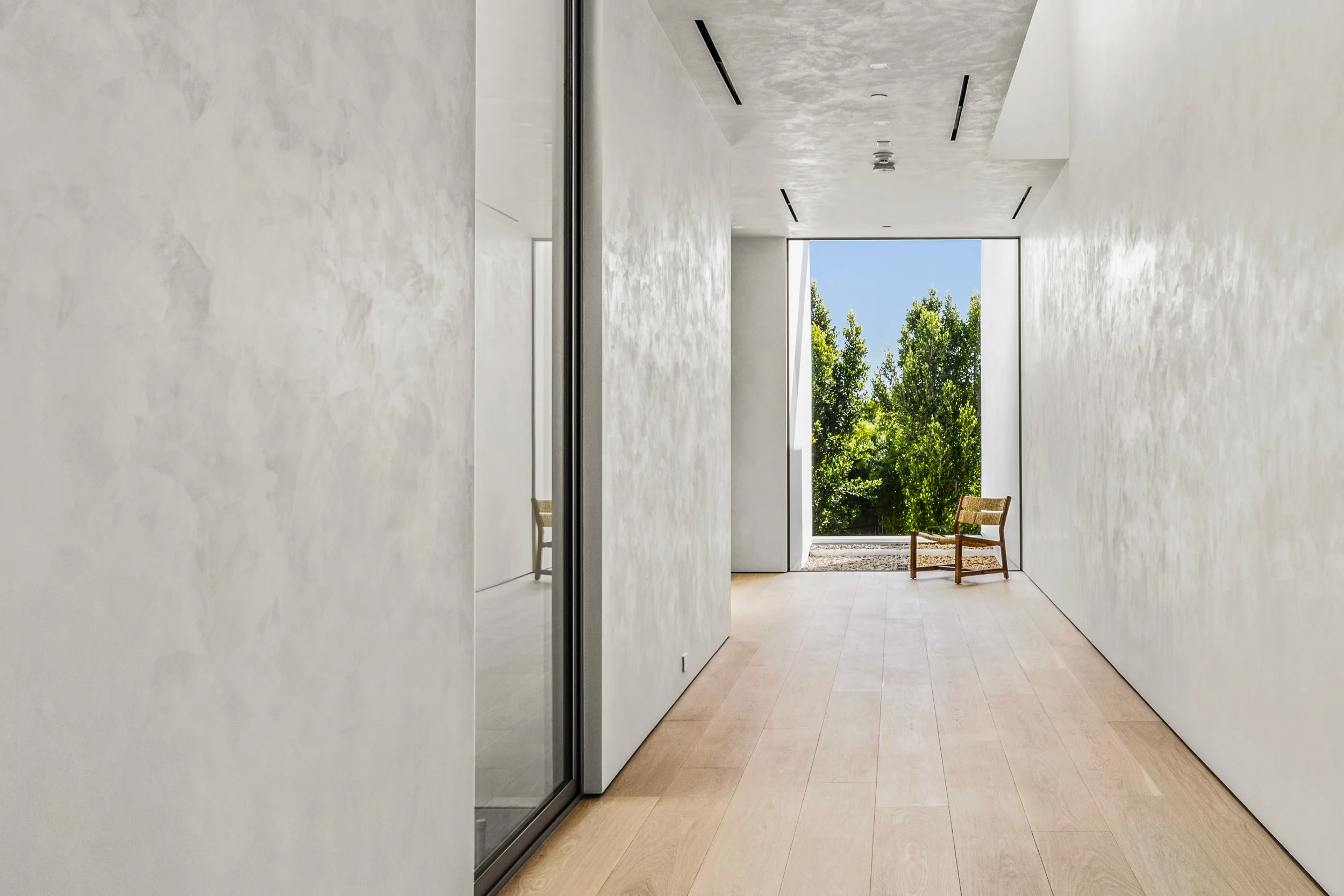 Minimal hallway with light wood floors, plaster walls, and a wooden chair by a sunlit window.