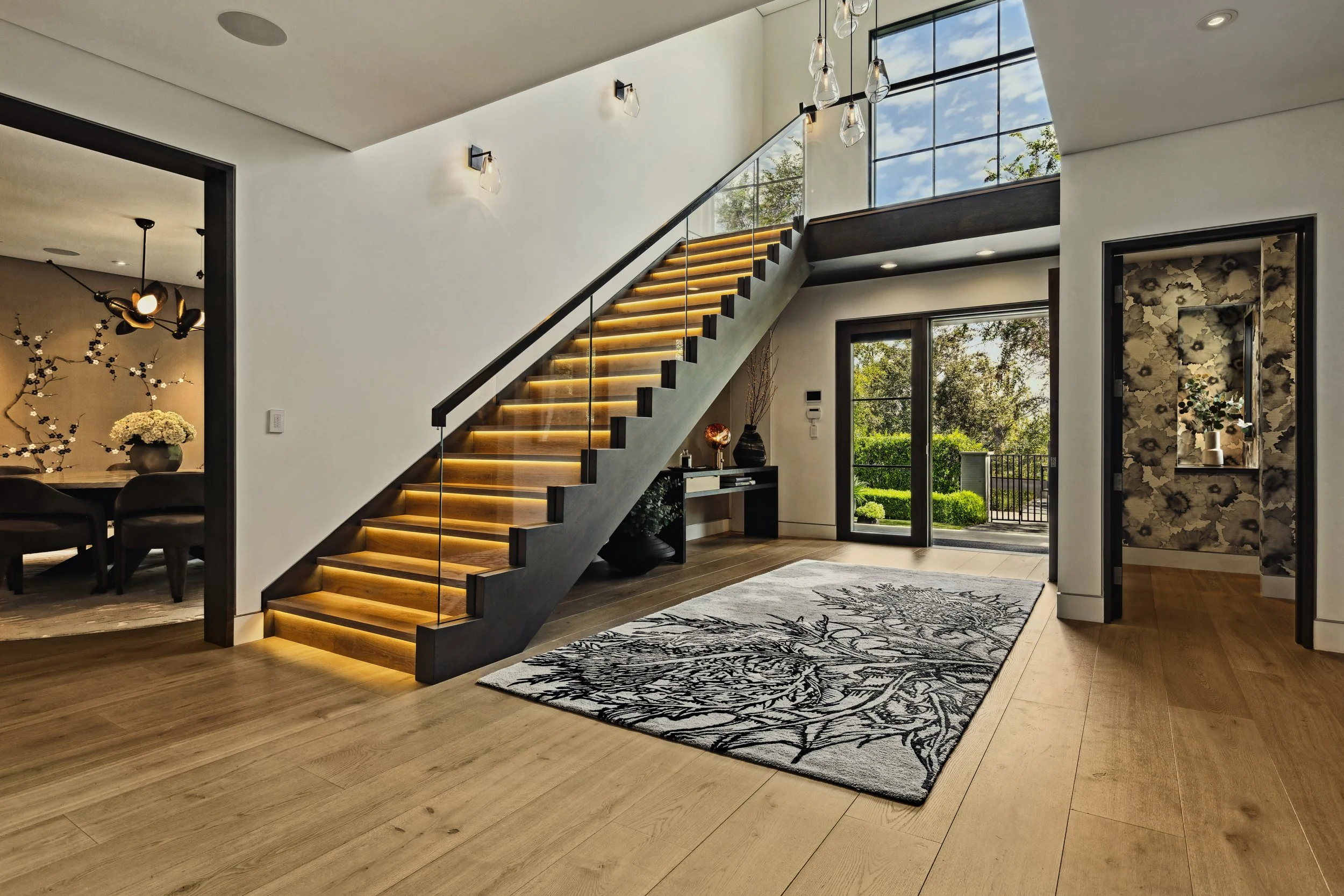 Foyer with a wood stair of lit treads, glass rails, and a bold black-and-white floral rug.