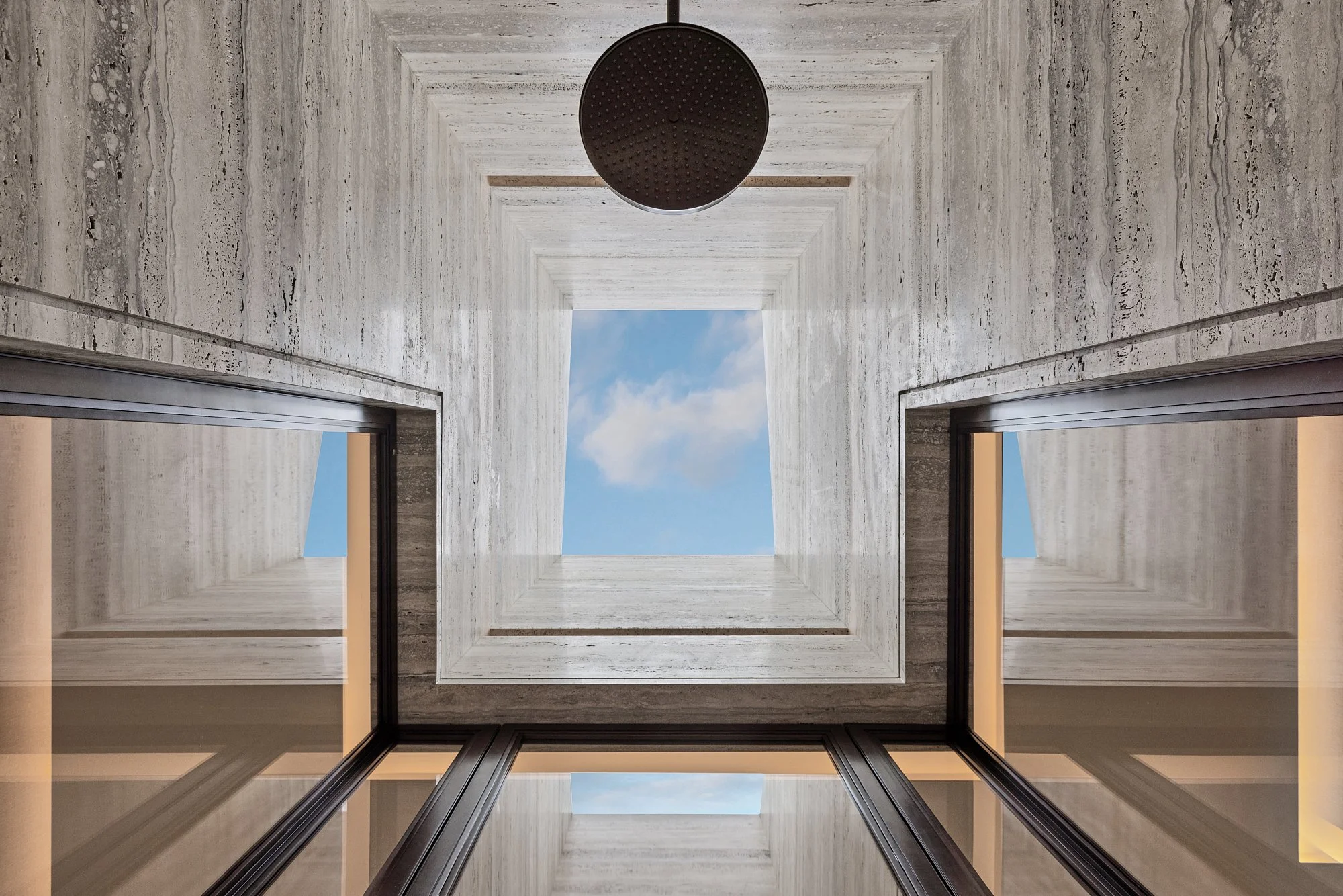 Upward view of a stone shower shaft with a black rain head open to a bright blue sky.