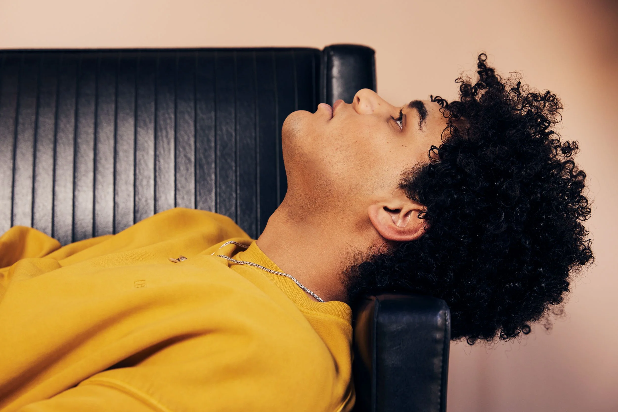 Portrait of Matthew Liner in a yellow sweatshirt resting his head back on a black leather sofa.