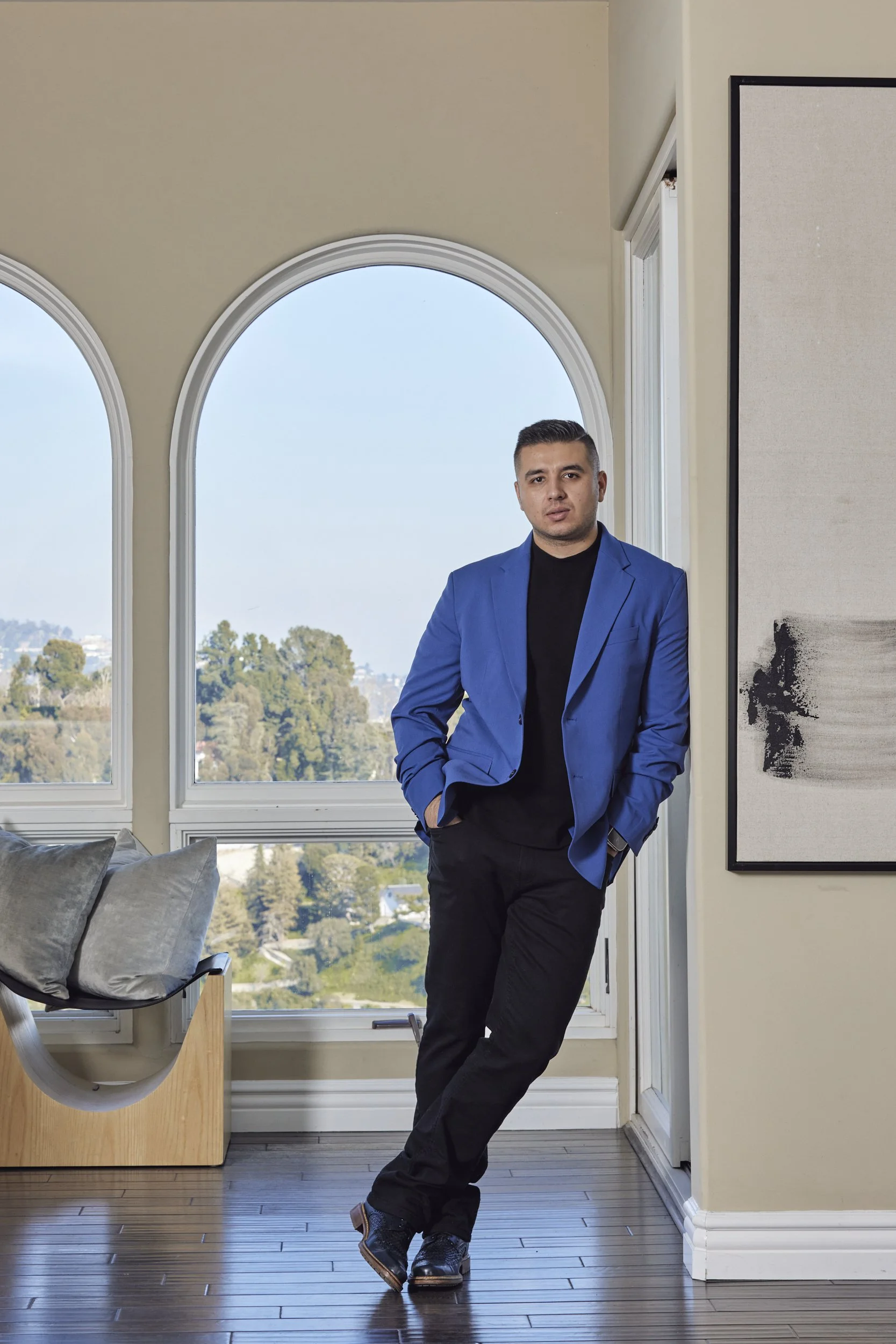 Portrait of Saul Ruvalcaba in a blue blazer leaning by arched windows overlooking a hillside.