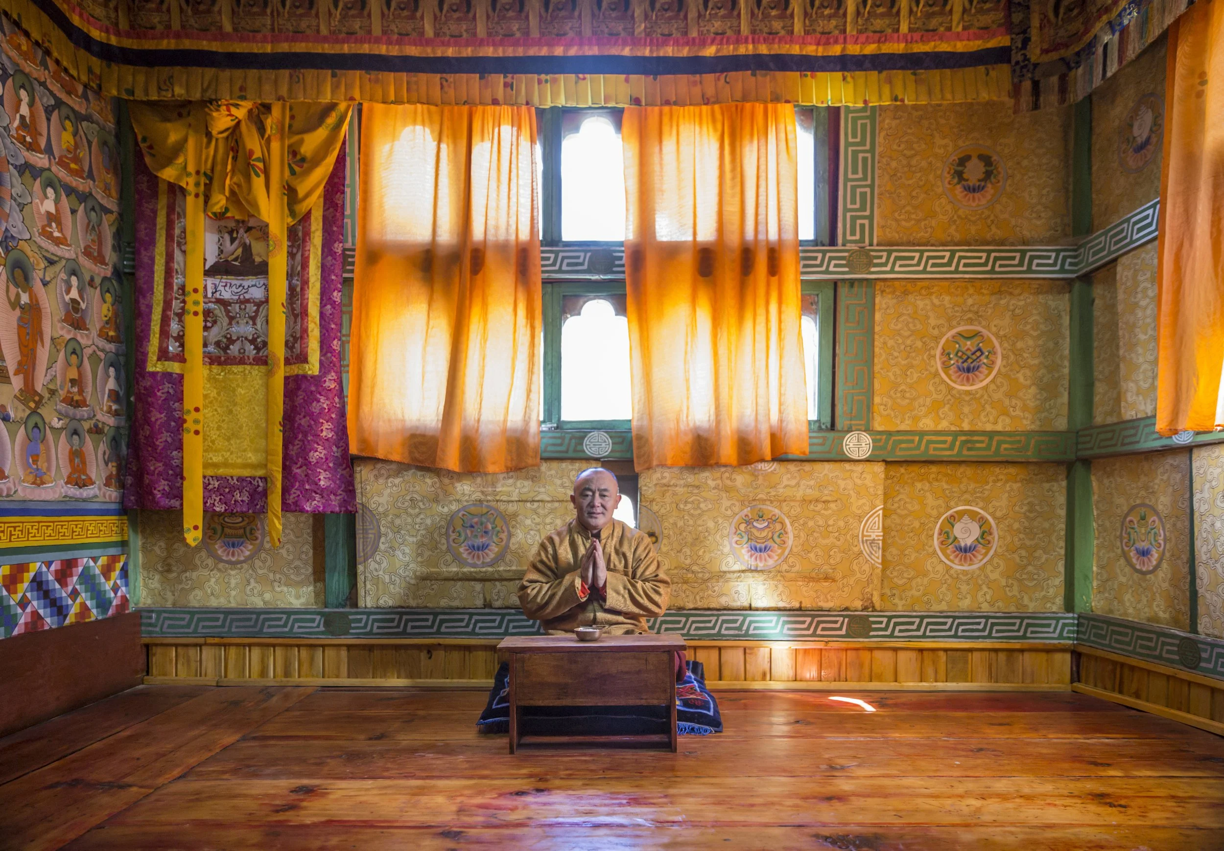 Man in yellow robes seated in prayer inside an ornate temple room lit by warm orange curtains.