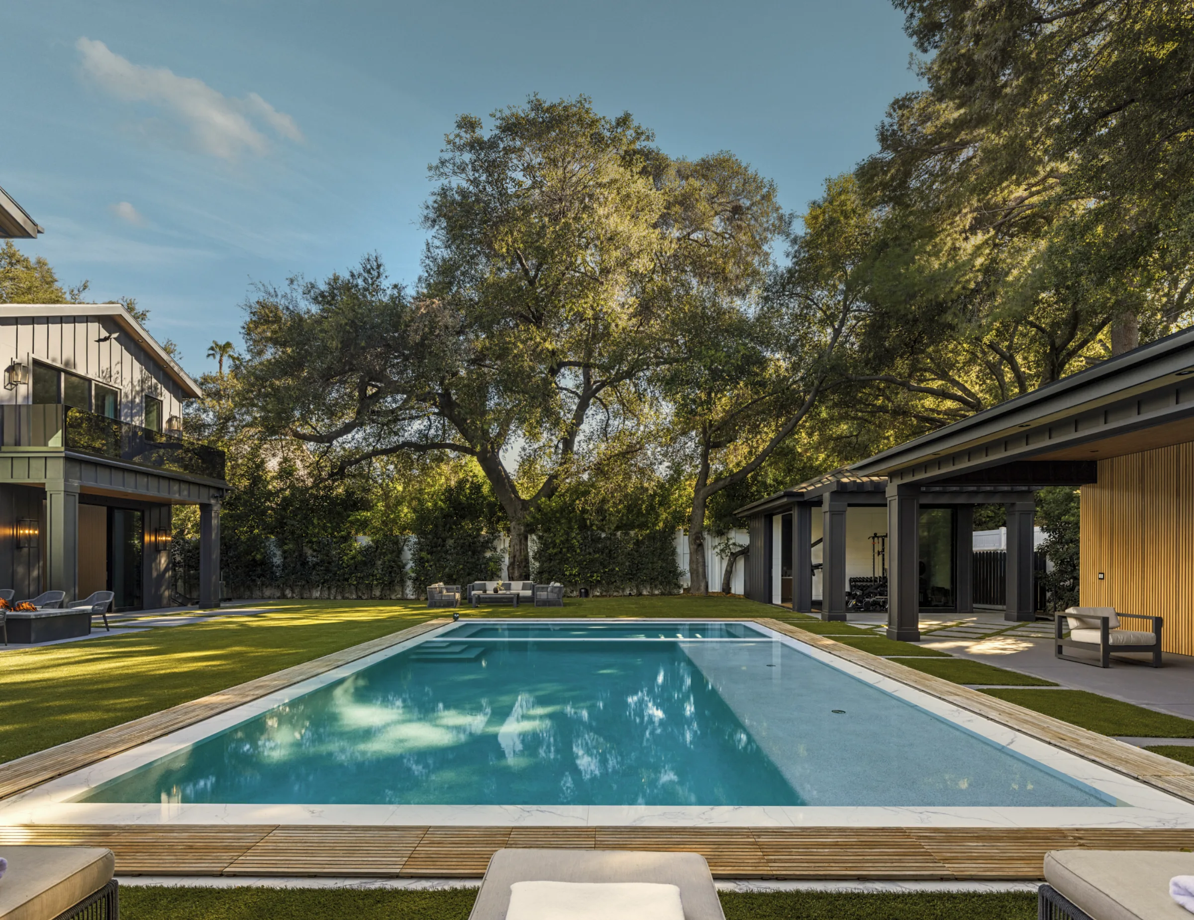 Zero-edge pool and wood deck before a dark-sided house and wood pavilion under oak trees.