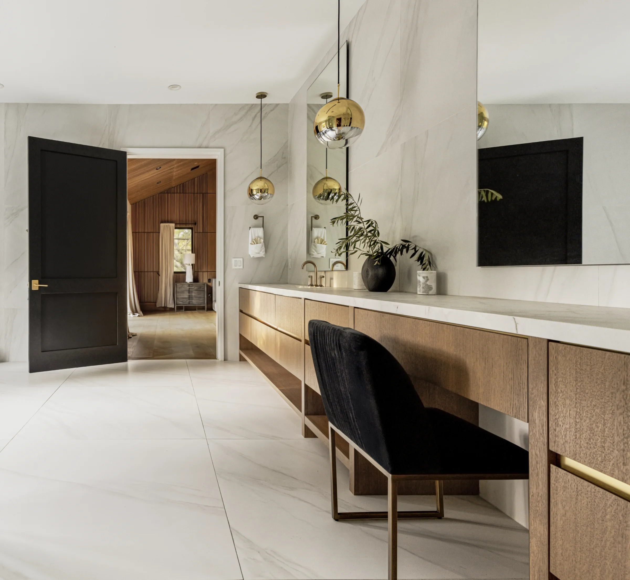 Long oak vanity with marble surfaces and gold pendants opening toward a wood-slat bedroom.
