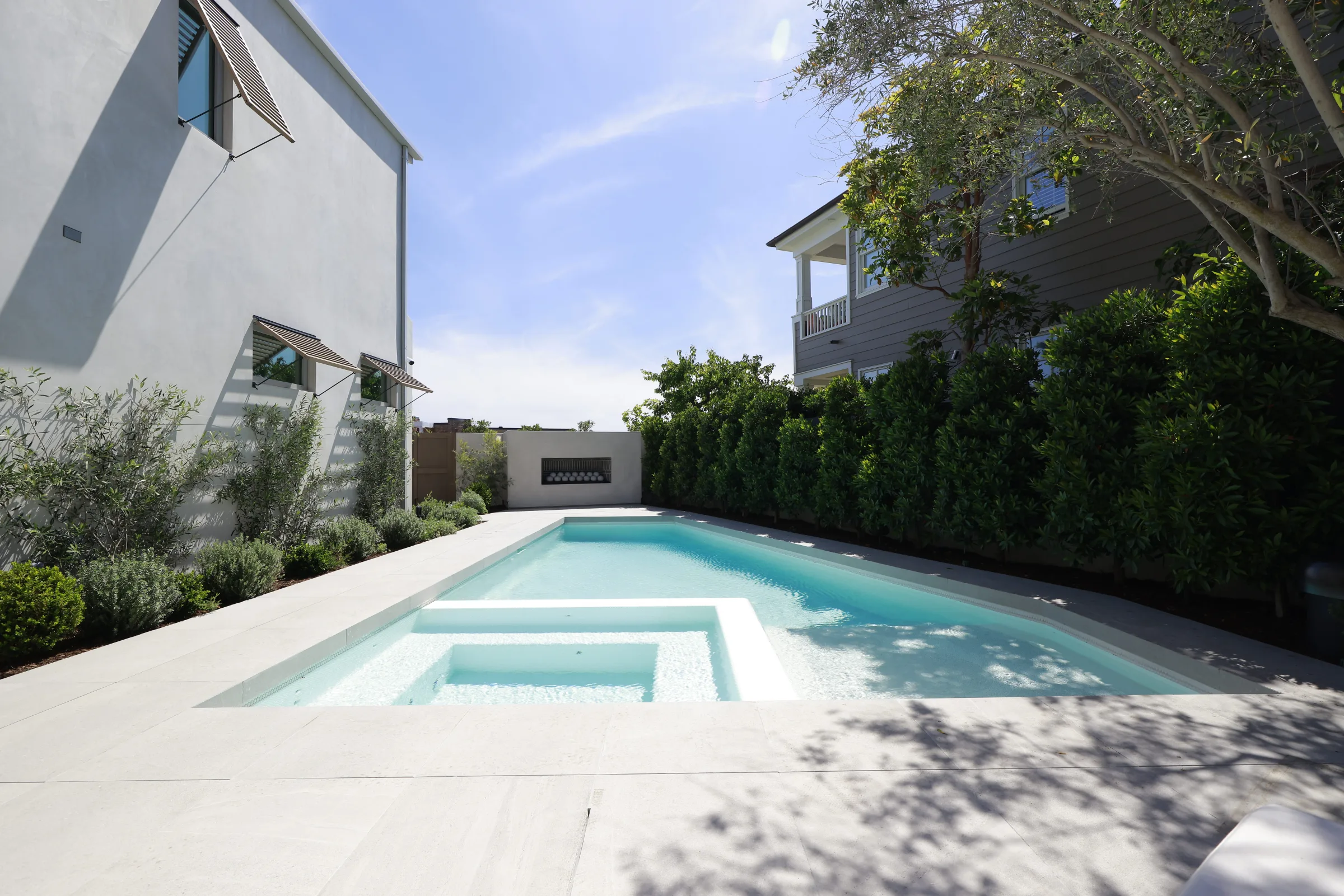 Long rectangular pool and spa on light stone decking between white walls and a privacy hedge.