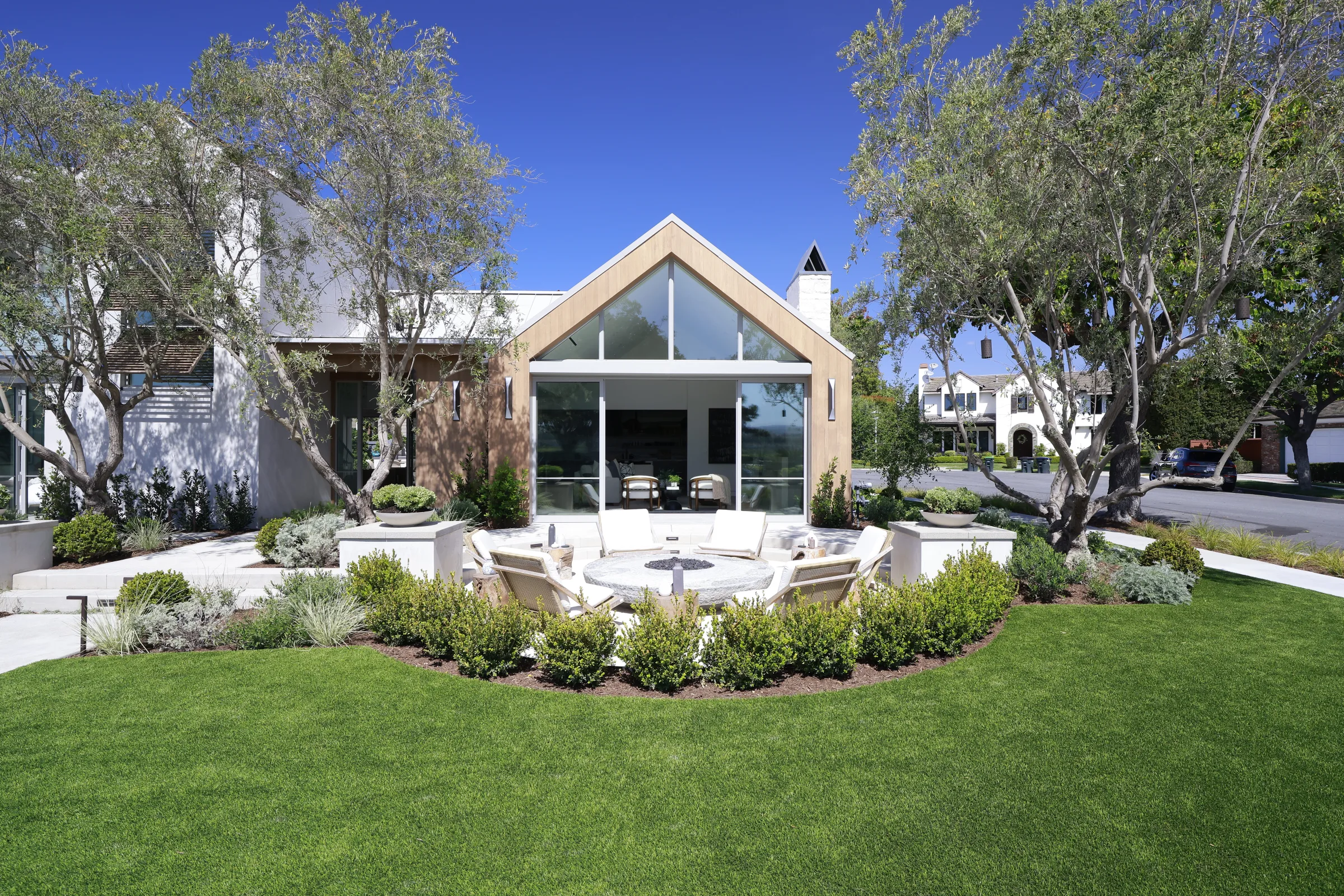 Wood-clad glass gable and circular fire lounge on a manicured lawn under a clear blue sky.