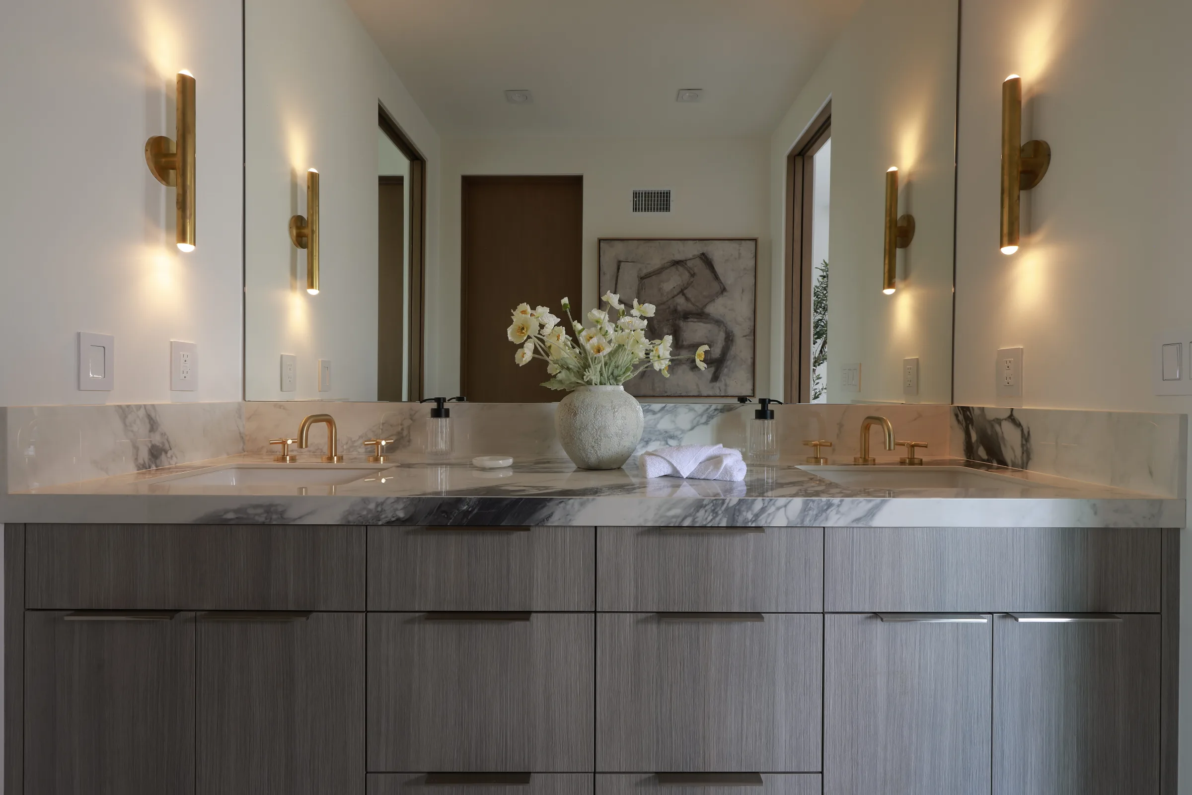 Double vanity with veined marble, light wood cabinets, and gold fixtures under soft sconces.